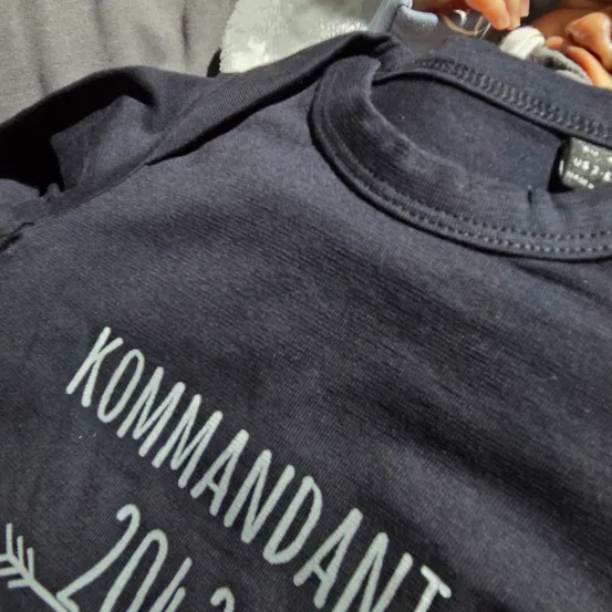 Ein schwarzes T-Shirt mit 'KOMMANDANT 2018' in Weiß auf der Vorderseite bedruckt. Eine Hand hält das Shirt.