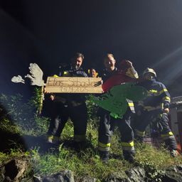 Feuerwehrleute stehen bei Nacht auf einem grasbewachsenen Hügel und halten ein Pappschild mit der Aufschrift 'Elias Sebastian 19.09.2025' mit Dinosaurier- und Vogelausschnitten. Sie tragen Helme und Uniformen.