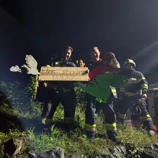 Feuerwehrleute stehen bei Nacht auf einem grasbewachsenen Hügel und halten ein Pappschild mit der Aufschrift 'Elias Sebastian 19.09.2025' mit Dinosaurier- und Vogelausschnitten. Sie tragen Helme und Uniformen.