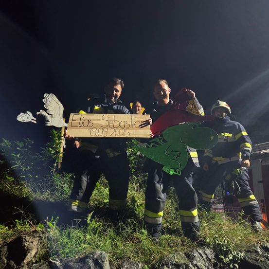 Feuerwehrleute stehen bei Nacht auf einem grasbewachsenen Hügel und halten ein Pappschild mit der Aufschrift 'Elias Sebastian 19.09.2025' mit Dinosaurier- und Vogelausschnitten. Sie tragen Helme und Uniformen.