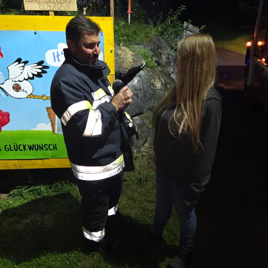 Ein Mann in Feuerwehruniform steht vor einer Frau und hält ein Mikrofon. Dahinter befindet sich ein gelbes Schild mit einem Storchbild und dem Wort 'Es'.