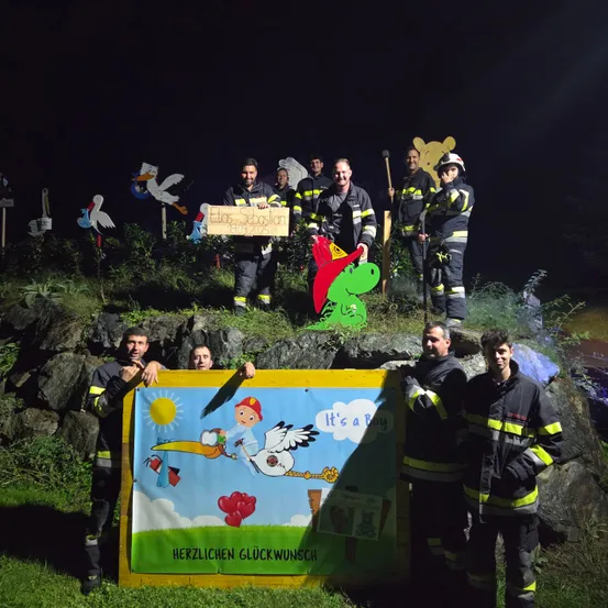 Feuerwehrleute posieren mit einem Banner und einem Schild, umgeben von dekorativen Elementen wie einem Storch und einem Frosch, unter einem Nachthimmel.