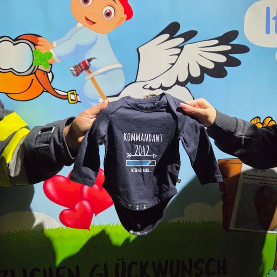 Zwei Hände halten den blauen Baby-Bodysuit mit der Aufschrift 'Kommandant 2042'. Im Hintergrund ist ein Cartoon-Feuerwehrmann, ein Vogel und Herzen zu sehen. Eine Sprechblase sagt 'Es.'