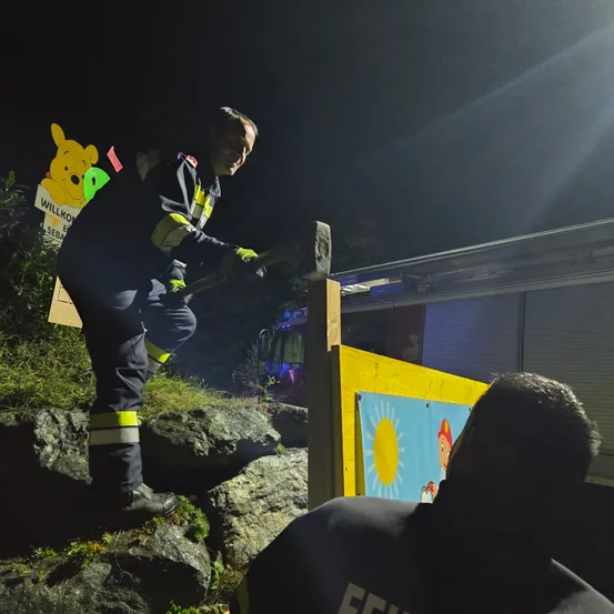 In der Nacht steht ein Feuerwehrmann auf einer felsigen Oberfläche und hält einen Hammer. Hinter ihm befindet sich ein Schild mit Winnie Puuh, das auf einer Holzstruktur platziert ist. Ein weiterer Feuerwehrmann betrachtet ein Schild mit einer Sonnenillustration. Ein Feuerwehrwagen ist auf der Seite geparkt.