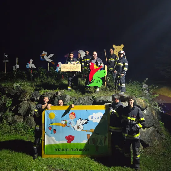 Feuerwehrleute posieren nachts mit einem Schild, Dekorationen und einem Storch. Das Schild sagt: 'Es ist ein Junge.' Sie halten einen Storch und ein Schild, auf dem steht: 'Alles Gute zum Geburtstag.'