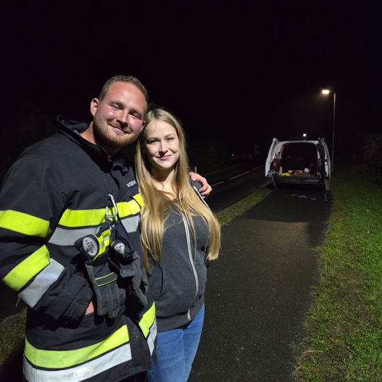 Ein Mann und eine Frau in Feuerwehrausrüstung stehen nachts am Straßenrand. Der Mann hat einen Helm und Handschuhe, und die Frau lächelt. Ein Feuerwehrwagen ist in der Nähe geparkt.
