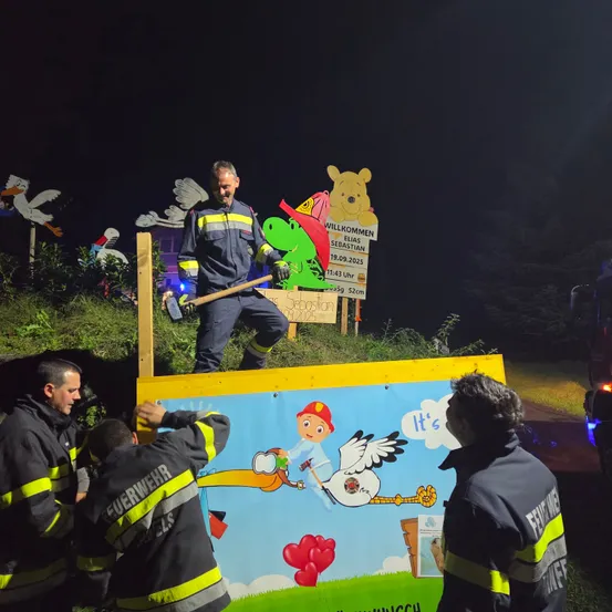 Ein Feuerwehrmann steht auf einer Holzplanke und hält einen Vorschlaghammer. Andere Feuerwehrleute halten ein Banner mit einem Storch und einem Herzen. Dahinter befindet sich ein Schild, das Elias Sebastian willkommen heißt.