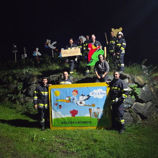 Eine Gruppe von Feuerwehrleuten posiert nachts für ein Foto, hält ein Schild mit der Aufschrift 'Es ist ein Junge' und 'Herzlichen Gluckwunsch'. Sie sind von Requisiten wie einem Storch, einem Schild und einigen Bäumen umgeben.