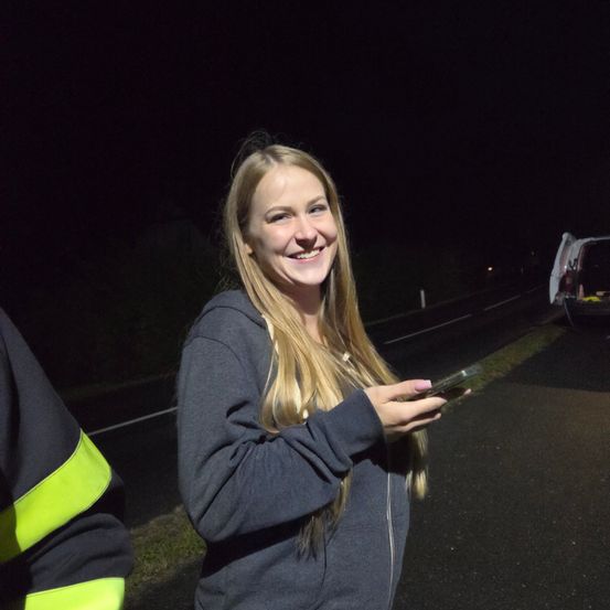Eine Frau mit blonden Haaren lächelt und schaut auf ihr Handy auf einer dunklen Straße bei Nacht.