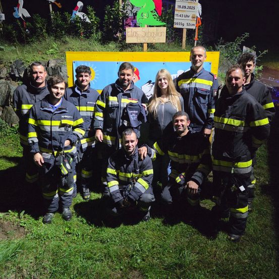 Eine Gruppe von Feuerwehrleuten posiert für ein Foto vor einem Schild. Das Schild trägt den Namen "Elas Sebasian" und hat ein Datum und eine Höhenangabe.