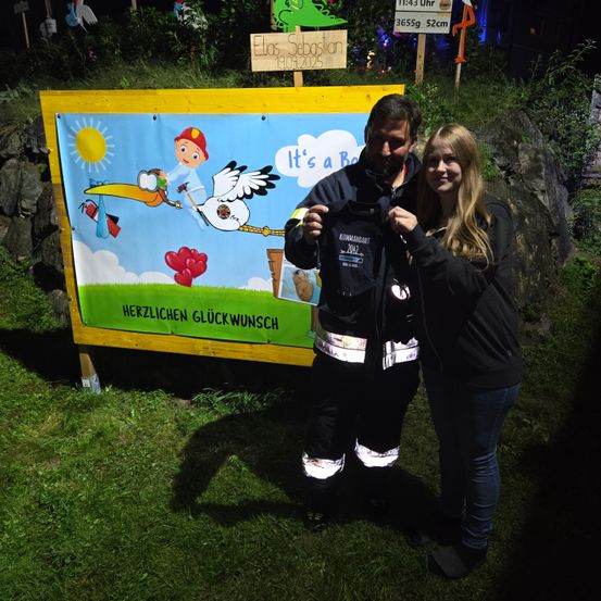 Ein Mann und eine Frau stehen vor einem bunten Schild, das 'Es ist ein Junge' sagt. Der Mann trägt eine reflektierende Jacke mit der Aufschrift 'KOMMANDANT 2022'. Sie lächeln und posieren für ein Foto. Dahinter ist ein Grasbereich mit einer Steinmauer.