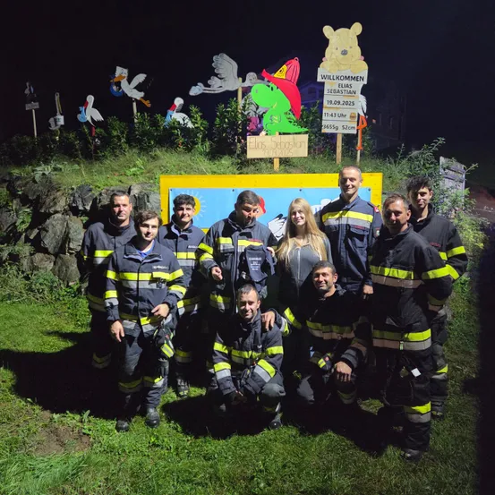 Eine Gruppe Feuerwehrleute in Uniform posiert nachts für ein Foto mit einem gelben Banner im Hintergrund. Dahinter befinden sich verschiedene Schilder und Dekorationsartikel.