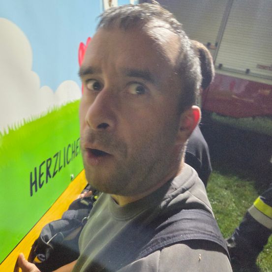 Ein Mann mit überraschtem Gesichtsausdruck schaut direkt in die Kamera. Hinter ihm steht ein grünes und gelbes Schild mit der Aufschrift Herzliche.