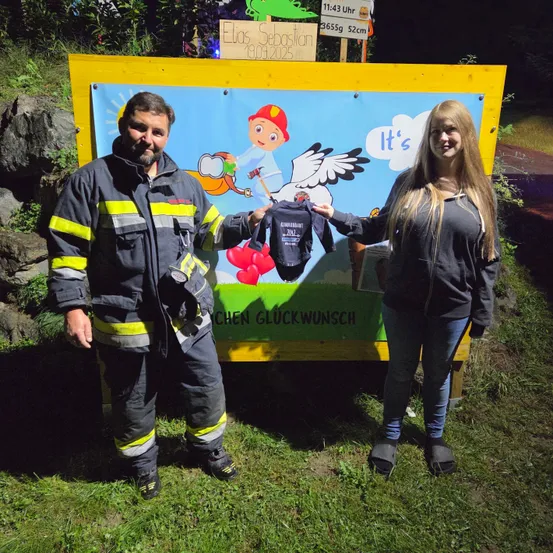 Ein Feuerwehrmann und eine Frau posieren mit einem Baby-Body draußen, in der Nähe eines Schildes, das 'Es ist ein Junge' sagt. Das Datum 19.06.2025 steht auch auf dem Schild.