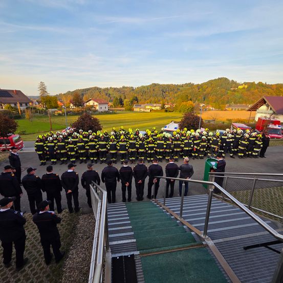 Eine Gruppe von Feuerwehrleuten steht in Formation auf einem Feld, umgeben von Feuerwehrwagen und Bergen in der Ferne.
