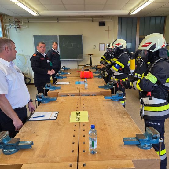 Mehrere Feuerwehrleute in voller Ausrüstung stehen in einem Raum mit einem Holztisch und diskutieren mit zwei Offizieren. Auf dem Tisch befinden sich verschiedene Gegenstände, darunter ein gelber Zettel und eine Wasserflasche.
