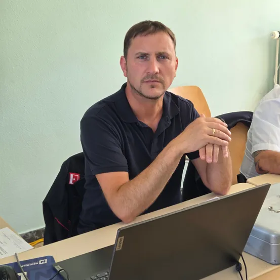 Ein Mann sitzt in einem Klassenzimmer an einem Schreibtisch, trägt ein schwarzes Poloshirt und hat einen Laptop und Kopfhörer in der Nähe. Er hat einen ernsten Gesichtsausdruck und stützt die Ellbogen auf dem Schreibtisch ab.