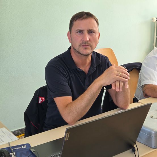 Ein Mann sitzt in einem Klassenzimmer an einem Schreibtisch, trägt ein schwarzes Poloshirt und hat einen Laptop und Kopfhörer in der Nähe. Er hat einen ernsten Gesichtsausdruck und stützt die Ellbogen auf dem Schreibtisch ab.