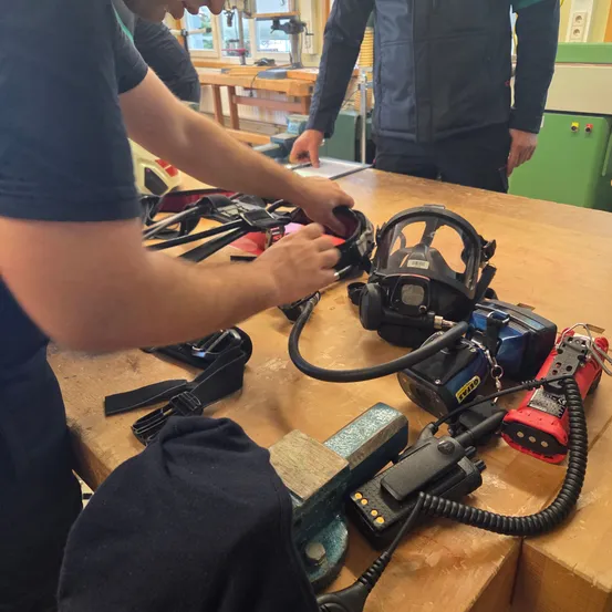 Zwei Personen arbeiten an einem Tisch mit Werkzeugen und Ausrüstung. Einer justiert eine Gasmaske, während ein anderer in der Nähe steht. Verschiedene Gegenstände, darunter ein Schleifer, liegen auf dem Tisch.