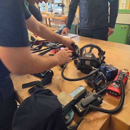 Zwei Personen arbeiten an einem Tisch mit Werkzeugen und Ausrüstung. Einer justiert eine Gasmaske, während ein anderer in der Nähe steht. Verschiedene Gegenstände, darunter ein Schleifer, liegen auf dem Tisch.