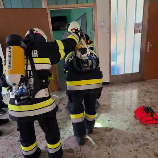 Zwei Feuerwehrleute in Schutzanzügen betreten einen Raum mit Glastür, einer trägt einen Feuerlöscher.