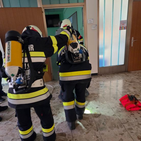 Zwei Feuerwehrleute in Schutzanzügen betreten einen Raum mit Glastür, einer trägt einen Feuerlöscher.