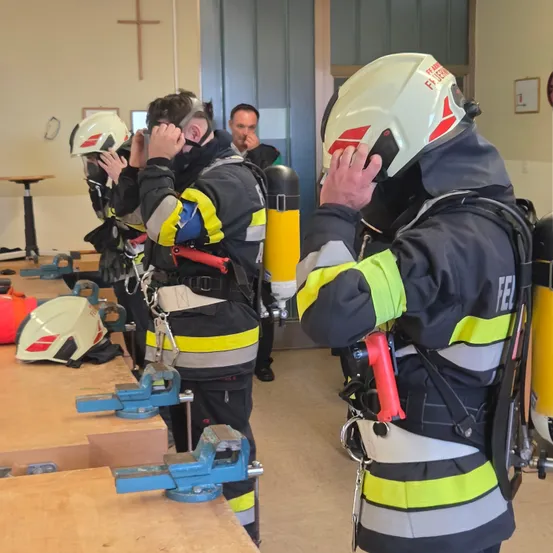 Mehrere Feuerwehrleute in Schutzausrüstung sind im Innenbereich. Ein Feuerwehrmann berührt seinen Helm, während andere beobachten. Verschiedene Werkzeuge und Geräte stehen auf den Holztischen.