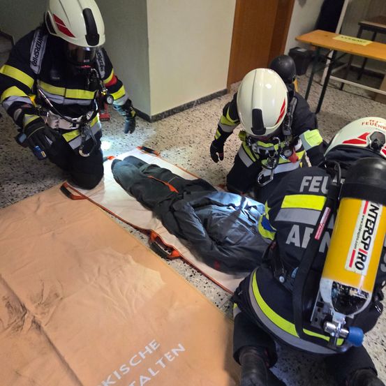 Vier Feuerwehrleute kümmern sich um eine Person auf einer Trage, die Schutzausrüstung tragen. Die Person ist in eine graue Plane gehüllt. Der Boden hat eine braune Matte mit den Worten 'KISCHE WEALTEN'. Ein Holztisch ist in der Nähe.