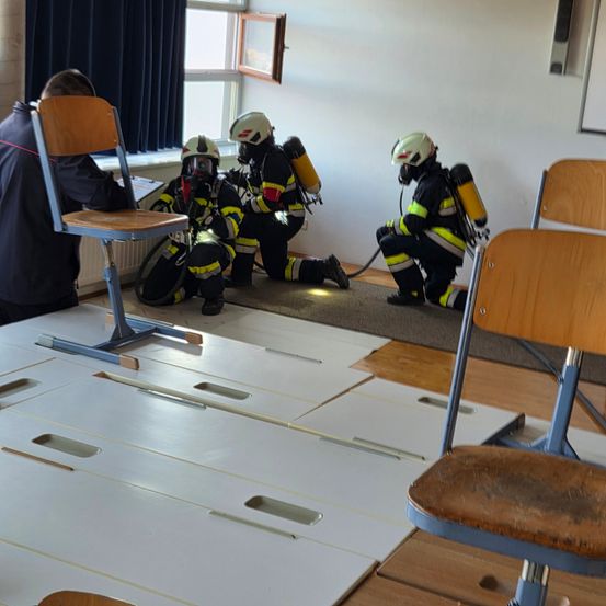 Drei Feuerwehrleute in voller Ausrüstung, einschließlich Sauerstoffflaschen, sind in einer Klassenzimmerumgebung und üben ihre Übungen auf dem Boden.