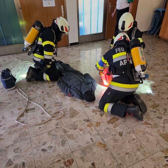 Feuerwehrleute in Schutzausrüstung retten eine Person auf dem Boden mithilfe einer Seile und Geräte. Die Rettungsaktion findet im Innenbereich in der Nähe einer Tür statt.