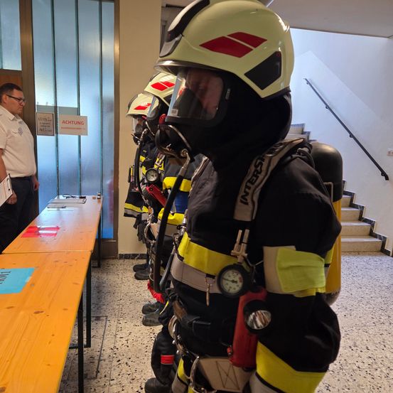 Eine Gruppe von Feuerwehrleuten in Schutzkleidung steht in einem Raum, einer hält einen roten Feuerlöscher. Ein Mann in einem weißen Hemd steht hinter ihnen und schaut auf einen Tisch mit Papieren.