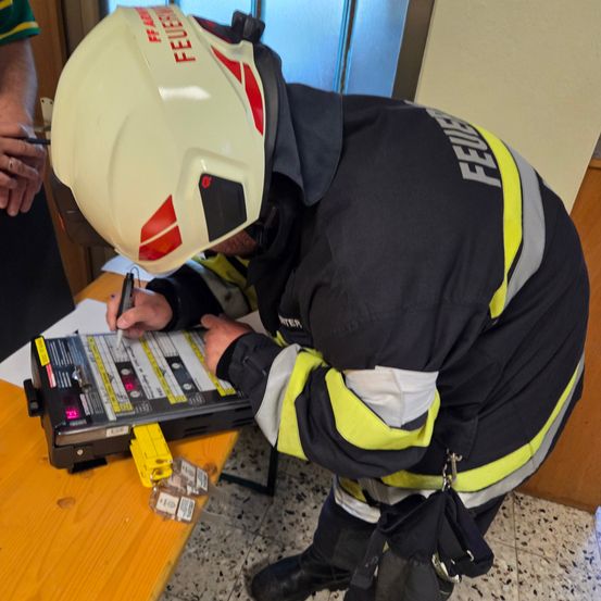 Ein Feuerwehrmann mit einem Helm, auf dem 'FEUER' steht, schreibt auf einem Klemmbrett auf einem Holztisch.