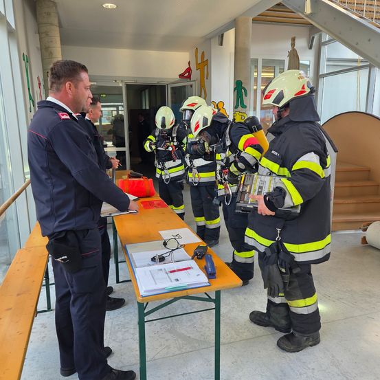 Feuerwehrleute in Schutzkleidung versammeln sich in einem Korridor und überprüfen Dokumente auf einem Tisch, während ein Mann in Uniform in der Nähe steht.