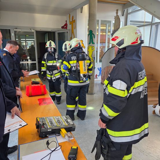 Feuerwehrleute in Schutzkleidung stehen in einem Gebäude und überprüfen Dokumente auf einem Tisch mit verschiedenen Geräten. Ein Mann im Vordergrund hält eine Clipboard.