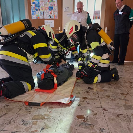 Feuerwehrleute in Schutzausrüstung üben eine Rettung an einem Dummy in einem Gebäude durch. Zwei knien, einer steht. Zwei Beobachter schauen von der Seite zu.