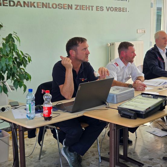 Drei Männer sitzen an einem Schreibtisch mit Laptops und Papieren. Der Mann auf der linken Seite schaut etwas an, während die anderen beiden ihm gegenübersitzen. Eine Pflanze, Flaschen und ein Heizkörper sind im Hintergrund.