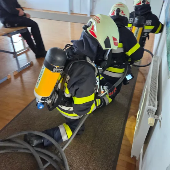 Ein Feuerwehrmann in voller Ausrüstung, einschließlich Helm und Sauerstoffflasche, liegt auf dem Boden und scheint an etwas zu arbeiten. Hinter ihm kniet ein weiterer Feuerwehrmann. In der Nähe befindet sich ein Heizkörper und eine Person, die auf einer Bank sitzt.