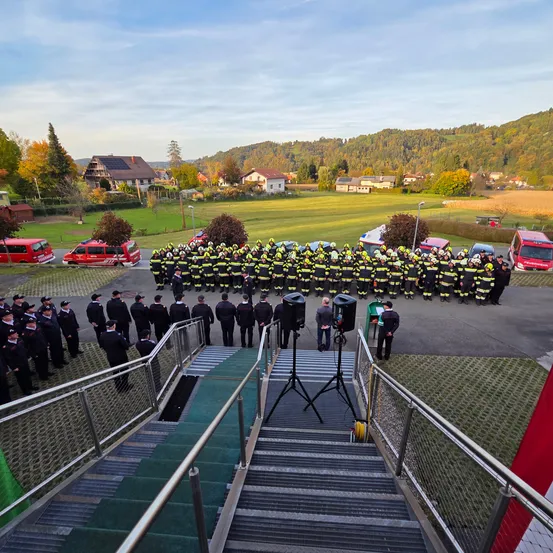 Eine Gruppe Feuerwehrleute in gelben und schwarzen Uniformen steht in Formation, mit roten Einsatzfahrzeugen hinter ihnen. Der Ort ist ein Feld mit Bergen in der Ferne.