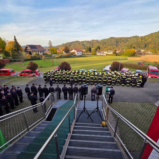 Eine Gruppe Feuerwehrleute in gelben und schwarzen Uniformen steht in Formation, mit roten Einsatzfahrzeugen hinter ihnen. Der Ort ist ein Feld mit Bergen in der Ferne.