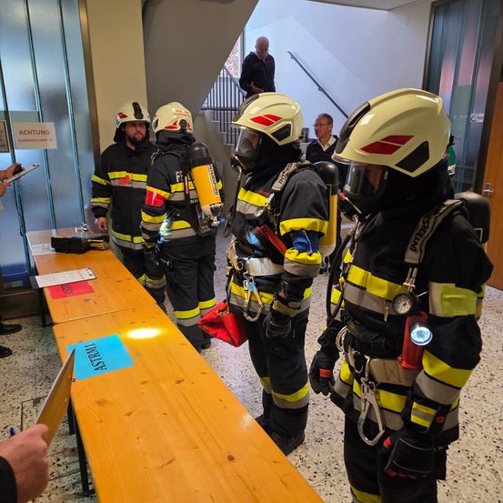 Fünf Feuerwehrleute in voller Ausrüstung stehen neben einem Tisch. Sie tragen gelbe und schwarze Uniformen, Helme und tragen Ausrüstung. Ein Mann steht auf einer Treppe hinter ihnen.
