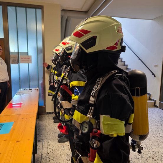 Feuerwehrleute in gelben und schwarzen Uniformen stehen in einem Flur vor einer Tür mit der Aufschrift 'Achtung'. Sie sind mit Helmen ausgestattet und tragen einen Feuerlöscher. Ein Mann in einem weißen Hemd steht neben der Tür.