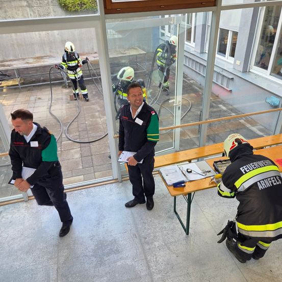 Eine Gruppe Feuerwehrleute in Uniform, einer mit einem Klemmbrett, stehen und knien in einem Glasraum. Draußen sind weitere Feuerwehrleute zu sehen.