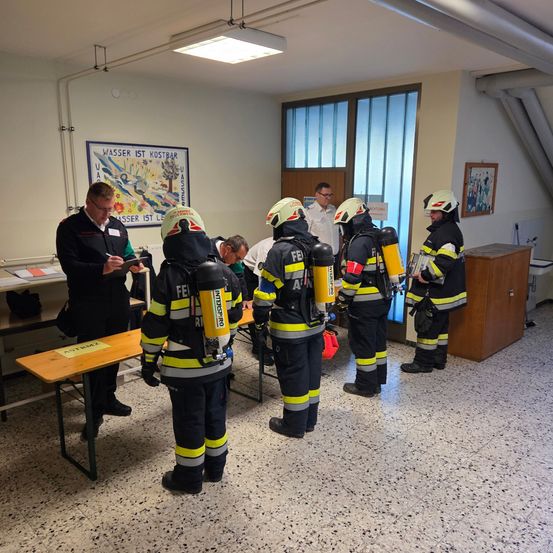 Mehrere Feuerwehrleute sind in einem Raum versammelt, wobei eine Person an einem Tisch sitzt. Die Feuerwehrleute tragen vollständige Ausrüstung, einschließlich Helme und Rucksäcke. Ein Plakat hängt an der Wand hinter ihnen.