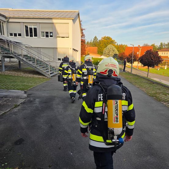 Feuerwehrleute in gelben Uniformen gehen auf einem Weg mit einem Gebäude und einer Treppe im Hintergrund. Sie tragen Ausrüstung und tragen Helme.