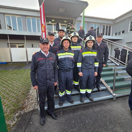 Eine Gruppe Feuerwehrleute, in Uniformen und Helmen gekleidet, steht auf den Stufen eines Gebäudes. Hinter ihnen ist eine Fahne zu sehen, und einige Personen stehen auf den Stufen.