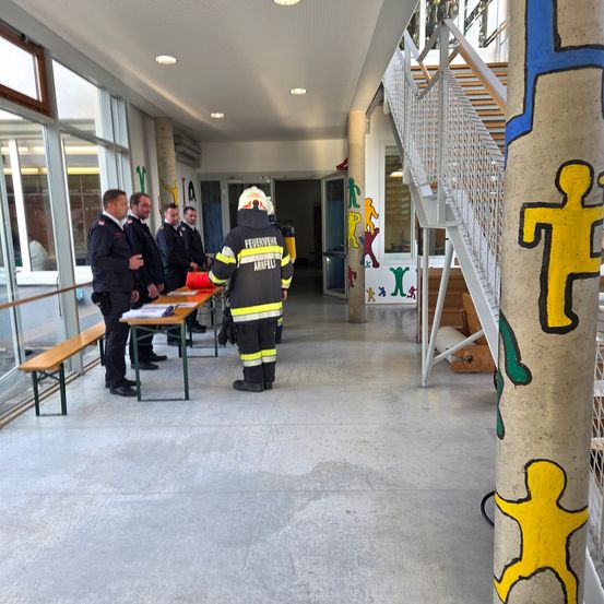 Mehrere Feuerwehrleute stehen in einem Korridor mit einem Tisch, während eine Person mit Helm mit ihnen spricht. In der Nähe befindet sich eine Treppe und ein Pfeiler mit bunten Figuren.