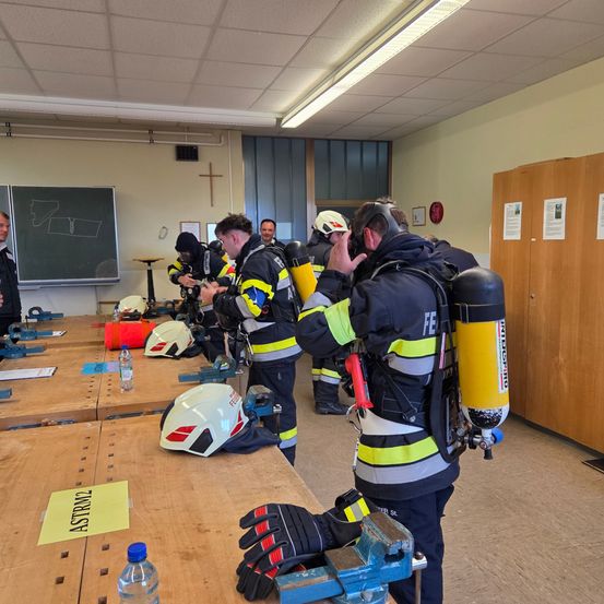 Eine Gruppe von Feuerwehrleuten in Schutzkleidung und Helmen steht in einem Klassenzimmer. Sie bereiten sich auf eine Übung vor. Einige passen ihre Ausrüstung an.