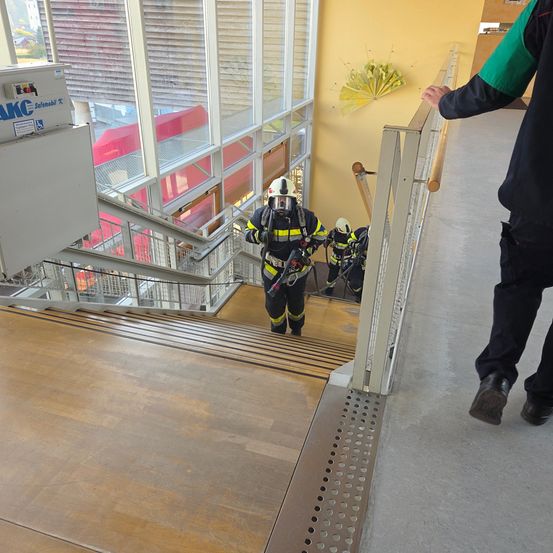 Drei Feuerwehrleute in Helmen und Schutzausrüstung steigen eine Treppe hinunter. Eine Person in schwarzen Hosen steht an der Seite und hält das Handgeländer. Glaswände und eine gelbe Wand sind sichtbar.