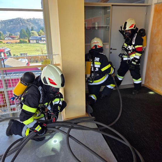 Drei Feuerwehrleute in voller Ausrüstung verlassen ein Gebäude mit einem Schlauch. Sie befinden sich in der Nähe einer Tür und einer Glaswand. Draußen gibt es eine Aussicht auf einen Berg und ein saftiges Feld.