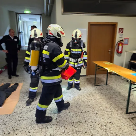 Feuerwehrleute in Schutzkleidung gehen in einem Raum. Einer hält ein rotes Objekt. Ein Mann in Anzug beobachtet. Ein Feuerlöscher ist an der Wand montiert.